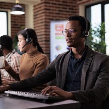 African american helpline employee working at call center reception with multiple monitors. Male operator using telecommunication to help clients at customer service support, remote network.