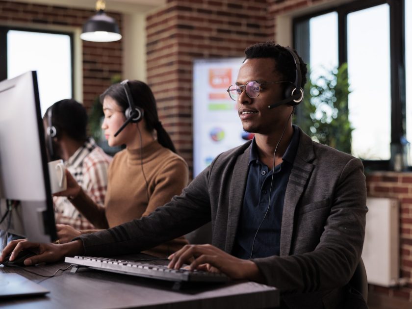 African american helpline employee working at call center reception with multiple monitors. Male operator using telecommunication to help clients at customer service support, remote network.